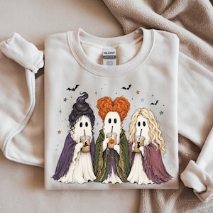 May include: A sand-colored crewneck sweatshirt featuring a graphic of three ghost figures dressed in long robes and distinct hairstyles, each holding a plastic cup with a straw. The design includes small bats and stars scattered above the ghosts.
