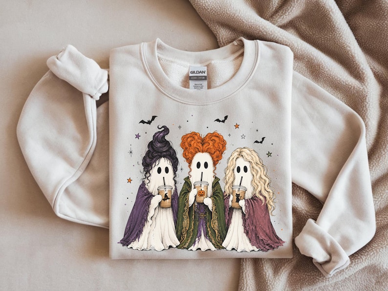 May include: A sand-colored crewneck sweatshirt featuring a graphic of three ghost figures dressed in long robes and distinct hairstyles, each holding a plastic cup with a straw. The design includes small bats and stars scattered above the ghosts.
