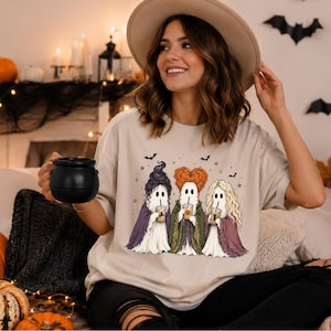 May include: A beige short-sleeve t-shirt featuring a graphic print of three ghost figures dressed as witches, each holding a coffee cup. The design includes small bats and stars, with the ghosts wearing purple, green, and pink cloaks.