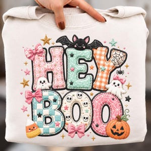 Op de afbeelding: Een crèmekleurig sweatshirt met de tekst HEY BOO in grote, gedessineerde letters. Het ontwerp bevat een zwarte vleermuis, witte spoken, een pompoen, maissnoepjes, schedels, roze strikken en kleine sterretjes in diverse kleuren.