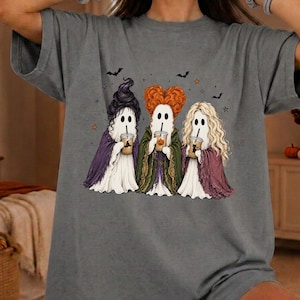 May include: A charcoal gray t-shirt featuring a graphic of three ghosts dressed as witches. Each ghost wears a different colored cape and holds a coffee cup. The ghosts have distinct hairstyles in black, orange, and blonde, with small bats and stars.