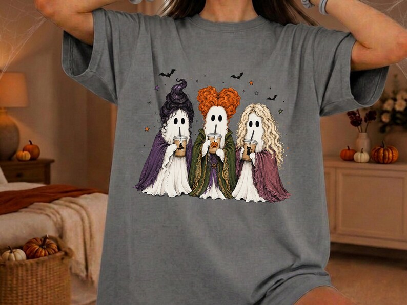 May include: A charcoal gray t-shirt featuring a graphic of three ghosts dressed as witches. Each ghost wears a different colored cape and holds a coffee cup. The ghosts have distinct hairstyles in black, orange, and blonde, with small bats and stars.