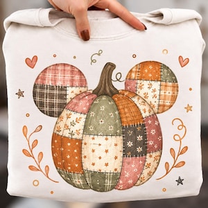 May include: A cream-colored sweatshirt featuring a large central graphic of a pumpkin with two smaller circular shapes above it, all designed with a patchwork pattern of floral, plaid, and solid autumn-toned fabrics in orange, green, and brown.