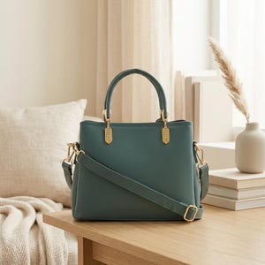 May include: A solid teal handbag featuring a structured rectangular body, two top handles, and a detachable shoulder strap. The bag is accented with gold-toned metal hardware, including rectangular connectors at the base of the handles.