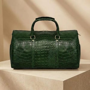 May include: A dark forest green holdall featuring a textured crocodile skin pattern. The bag has two top handles, gold-toned metal hardware, and a zipped closure along the top. The rectangular structure is consistent with a travel bag design.
