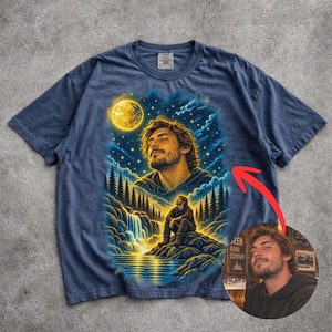 May include: A navy blue t-shirt featuring a graphic print of a person with wavy hair looking up at a bright yellow moon in a starry night sky. Below, the same person sits on a rock near a waterfall and pine trees. The design uses blue and yellow tones.