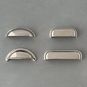 May include: Four polished silver-toned metal drawer pulls arranged in two pairs. The left pair features two semi-circular cup-style handles, while the right pair consists of two rectangular-shaped handles with rounded edges and a smooth finish.