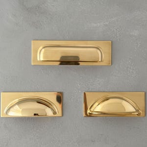 May include: Three rectangular brass drawer pulls with a polished finish. One large pull is positioned horizontally at the top, while two smaller, semi-circular cup-style pulls are arranged side-by-side below it, all featuring a smooth metallic surface.