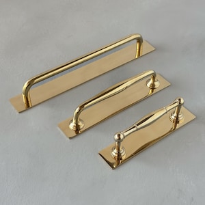 Op de afbeelding: Drie goudkleurige metalen kastgrepen, elk voorzien van een rechthoekige achterplaat. De bovenste greep heeft een eenvoudige buisvormige handgreep, de middelste is licht gebogen en de onderste greep heeft een decoratief ontwerp met ronde uiteinden.