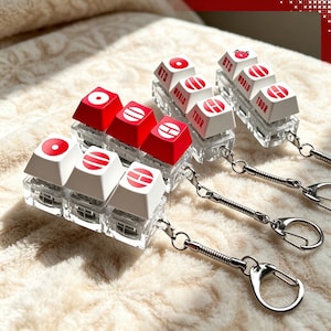 May include: A set of four mechanical keyboard switch keychains featuring clear acrylic bases and metal rings. The keycaps are white and red, displaying circular symbols and the text BTS WORLD TOUR, with one keycap featuring a small red cartoon character.