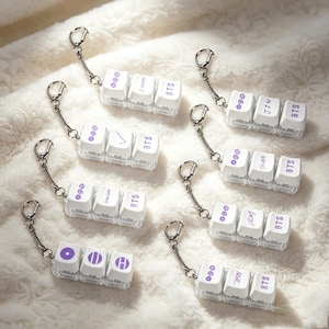 Könnte beinhalten: Eine Sammlung von acht Schlüsselanhängern, die jeweils aus drei weißen mechanischen Tastaturkappen an einem silbernen Metallring bestehen. Die Kappen zeigen violette Aufdrucke und Symbole, darunter BTS und Namen von Mitgliedern.