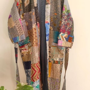 Könnte beinhalten: Ein langärmeliger Morgenmantel mit einem Patchwork-Design aus verschiedenen Stoffquadraten in unterschiedlichen Farben und Mustern. Das Kleidungsstück verfügt über einen passenden Bindegürtel, eine offene Vorderseite und sichtbare weiße Steppnähte.