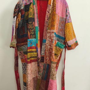 Op de afbeelding: Een kamerjas met lange mouwen en een patchwork-ontwerp bestaande uit diverse kleurrijke stoffen vierkanten met uiteenlopende bloem- en geometrische patronen. Het kledingstuk heeft een felrode binnenvoering en een bijpassende ceintuur.