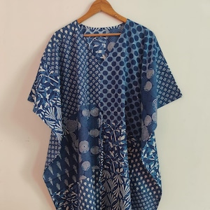 Può includere: Un caftano blu e bianco caratterizzato da un design patchwork con vari motivi geometrici e floreali, tra cui pois, cerchi concentrici e disegni di foglie. L'indumento presenta uno scollo a V, maniche corte ed è appeso a una gruccia in legno.