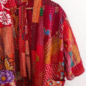 Könnte beinhalten: Eine langärmelige Jacke mit einem lebendigen Patchwork-Design in Rot-, Orange- und Violetttönen. Das Kleidungsstück besteht aus verschiedenen Stoffquadraten mit detaillierten weißen Steppstichen und verfügt über eine Vordertasche.