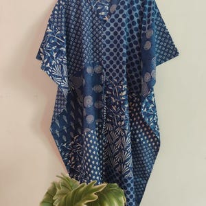 May include: A blue and white kaftan featuring a patchwork design of various geometric and floral patterns. The garment has a V-neckline, short sleeves, and a loose, flowing silhouette, displayed on a wooden hanger against a plain wall.