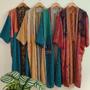 May include: Four long, open-front kimonos hanging on wooden hangers. Each garment features a unique combination of colorful, patterned fabric panels in shades of teal, orange, yellow, and purple, with long sleeves and a loose, flowing silhouette.