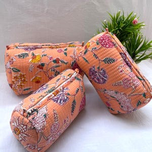 Puede incluir: Tres estuches cilíndricos acolchados de color melocotón suave, cada uno con un patrón floral detallado con acentos en morado, amarillo y verde. Los estuches tienen un cierre de cremallera y una superficie de tela texturizada y pespunteada.