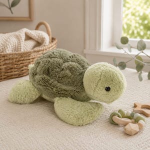 Könnte beinhalten: Ein Plüsch-Schildkrötenspielzeug mit einem weichen, strukturierten olivgrünen Panzer sowie einem Kopf und Flossen in einem helleren Salbeigrün. Das Stofftier hat einen abgerundeten Körper, kleine dunkle Augen und eine flauschige Oberfläche.