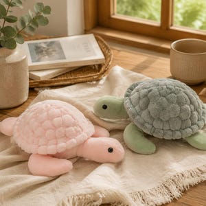 Puede incluir: Dos tortugas de peluche, una en rosa pastel suave y la otra en verde salvia apagado, con caparazones redondeados y texturizados y extremidades lisas. Cada tortuga tiene una cabeza redondeada sencilla con ojos negros pequeños, de tela suave.