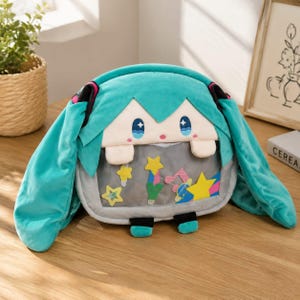 May include: A plush bag featuring a teal-haired character with long side ponytails, blue eyes, and pink cheeks. The front has a clear window pocket containing colorful felt shapes including stars, a planet, and musical notes, with two small teal feet.