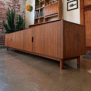 May include: A long, low-profile wooden sideboard featuring a rich brown finish and vertical slat detailing across the front doors. The cabinet is supported by four cylindrical legs and includes recessed, diamond-shaped handles on the sliding doors.