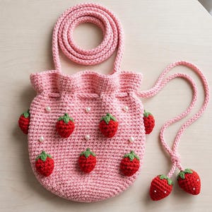Puede incluir: Un bolso de ganchillo rosa claro con cordón, decorado con pequeñas aplicaciones de fresas rojas con hojas verdes y perlas blancas dispersas. El bolso tiene una correa larga trenzada y dos colgantes de fresa en los extremos.