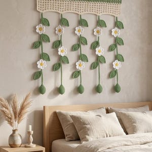 May include: A crochet wall hanging featuring five vertical strands of green stems with white flowers and yellow centers, suspended from a wooden rod. The top section consists of a green and cream-colored grid pattern with loops for hanging.