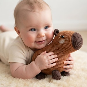 May include: A brown crocheted capybara soft toy with a dark brown snout, small rounded ears, and black bead eyes. The toy features a textured yarn construction and is shaped like a capybara, showcasing a rounded body and four small feet.