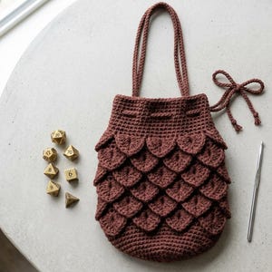 May include: A dark brown crochet drawstring bag featuring a textured scale pattern on the exterior. The bag includes two long handles and a matching cord tie. Beside the bag are six gold-colored polyhedral dice and a silver metal crochet hook.