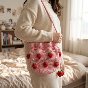 Puede incluir: Un bolso de ganchillo rosa claro con cierre de cordón, decorado con pequeñas aplicaciones de fresas rojas con hojas verdes y detalles de perlas blancas. El bolso incluye una correa trenzada rosa y dos colgantes de fresa en el cordón.