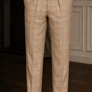 May include: Tan trousers featuring a subtle blue windowpane check pattern, double pleats at the waist, and side adjusters with silver-toned buckles. The trousers have a straight-leg cut with cuffed hems and are paired with a light-colored shirt.