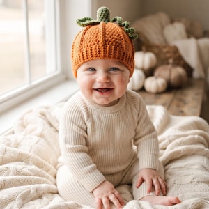 May include: A crocheted orange pumpkin hat with a green stem and leaf detail on top. The hat features a ribbed texture and a rounded shape, designed to cover the head with a snug fit, showcasing detailed yarn work and a vibrant orange color.