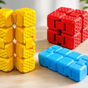 Può includere: Tre giocattoli antistress articolati composti da cubi in plastica. Uno è giallo brillante e posizionato verticalmente, uno è rosso e ripiegato in un blocco compatto, e uno è azzurro e appoggiato in piano. Ogni cubo ha una trama a griglia.