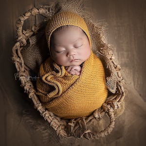 Op de afbeelding: Een pasgeboren baby slaapt rustig in een gevlochten mand, gewikkeld in een zacht, geel gebreid dekentje en mutsje. De baby draagt een bijpassend gebreid mutsje.