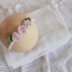 以下が含まれることがあります： 白いヘッドバンドに、ピンクの布の花が3つとパールがあしらわれています。ヘッドバンドは木製のボールと白い編み物の毛布の上に置かれています。