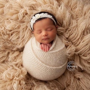 Op de afbeelding: Een pasgeboren baby gewikkeld in een witte doek, slaapt op een lichtbruin, pluche deken. De baby draagt ​​een wit hoofdband met bloemen.