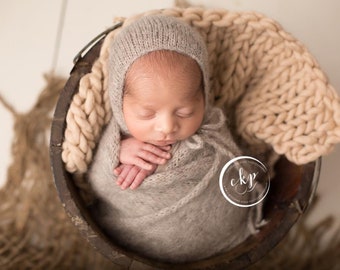 May include: A newborn baby is sleeping soundly in a wooden bucket, wrapped in a gray knitted blanket and wearing a matching knitted hat. The baby is surrounded by a beige knitted blanket.