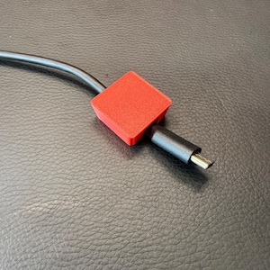May include: A bright red, square-shaped cable protector clip attached to a black charging cable. The accessory features a textured surface and is positioned around the cable near the metallic connector end, providing a secure fit for the cord.