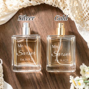 May include: Two square glass perfume bottles with spray nozzles. The left bottle has a silver cap and is engraved with "Mr. Sarver Est. 16.9.25". The right bottle has a gold cap and is engraved with "Mrs. Sarver Est. 16.9.25". Both contain amber liquid.