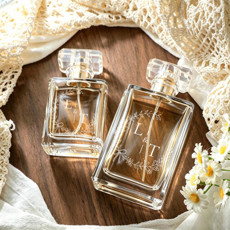 May include: Two rectangular glass perfume bottles with clear caps and gold-toned spray nozzles. Each bottle features an etched white floral wreath design surrounding the letters L & T. The bottles contain a light amber liquid and have clear glass.