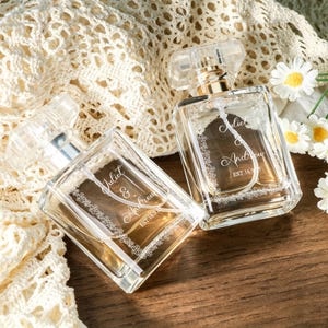 May include: Two clear glass perfume bottles with rectangular shapes and transparent caps. Each bottle features a white floral border engraving and the text Juliet & Andrew EST. 16.9.25. The bottles are positioned on a wooden surface.