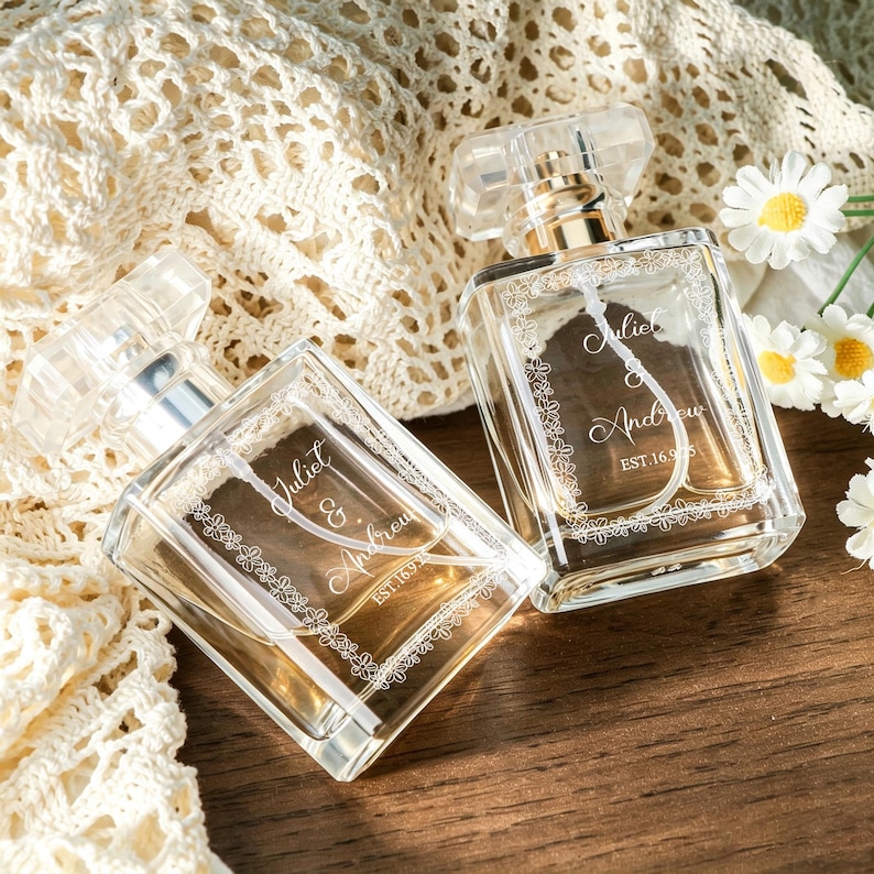 May include: Two clear glass perfume bottles with rectangular shapes and transparent caps. Each bottle features a white floral border engraving and the text Juliet & Andrew EST. 16.9.25. The bottles are positioned on a wooden surface.