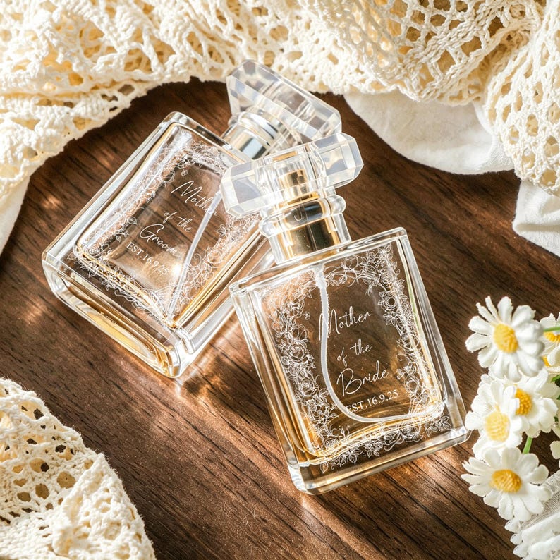May include: Two clear glass perfume bottles with rectangular shapes and transparent caps. One bottle is engraved with Mother of the Groom EST.16.9.25 and the other with Mother of the Bride EST.16.9.25, both featuring intricate floral border designs.