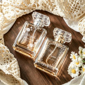 May include: Two rectangular glass perfume bottles with clear caps and gold-toned spray mechanisms. The left bottle is engraved with 'Angela BRIDESMAID EST. 16.9.2025' and floral accents. The right bottle features 'Mr. Sarver 16.9.2025' with a floral border.