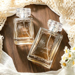 May include: Two rectangular glass perfume bottles with clear caps, each featuring an etched floral vine design and the text "Mrs. Sarver 16.9.2025" on the front. The bottles contain a light amber liquid and have a transparent spray nozzle inside.