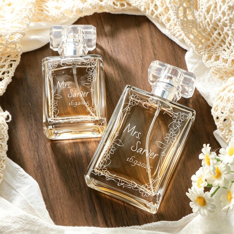 May include: Two rectangular glass perfume bottles with clear caps, each featuring an etched floral vine design and the text "Mrs. Sarver 16.9.2025" on the front. The bottles contain a light amber liquid and have a transparent spray nozzle inside.
