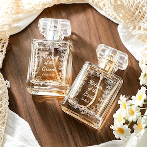 May include: Two clear glass perfume bottles with rectangular bodies and transparent caps. Each bottle features an etched floral border and white text. One reads Mother of the Groom Est. 16.9.25, and the other reads Mother of the Bride Est. 16.9.25.