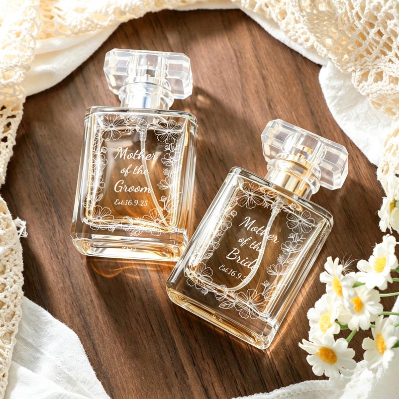 May include: Two clear glass perfume bottles with rectangular bodies and transparent caps. Each bottle features an etched floral border and white text. One reads Mother of the Groom Est. 16.9.25, and the other reads Mother of the Bride Est. 16.9.25.