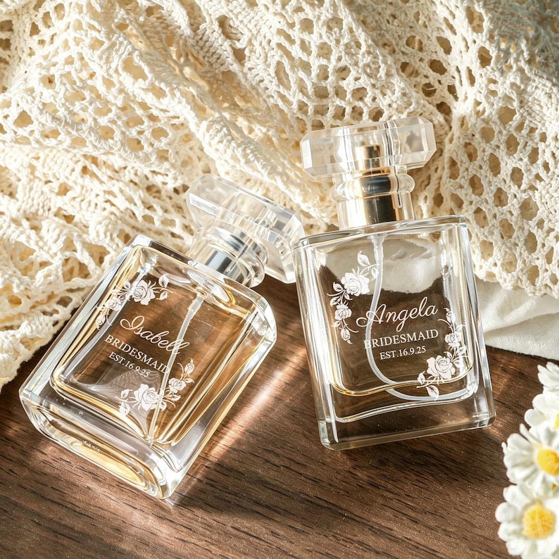 May include: Two clear glass perfume bottles with rectangular shapes and transparent caps. Each bottle features white etched floral designs and text reading Isabell or Angela, Bridesmaid, and EST.16.9.25, filled with a light amber liquid.